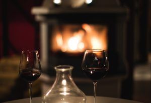 Two glasses of red wine sit on either side of a decanter on a table in front of a glowing electric fireplace.
